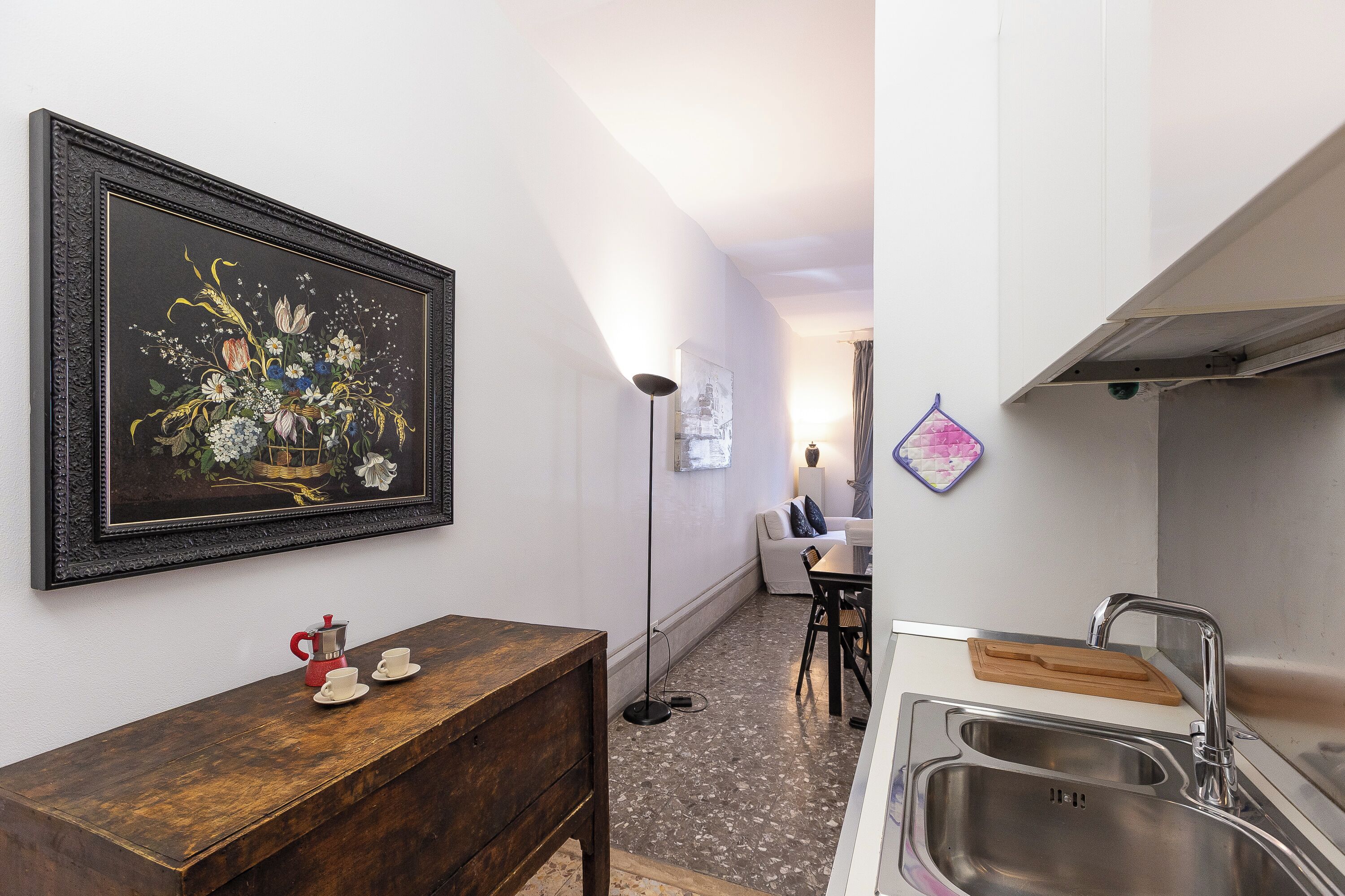 Una cucina con un lavello, un tavolo di legno con una macchina del caffè rossa, un quadro floreale incorniciato e una lampada da terra.