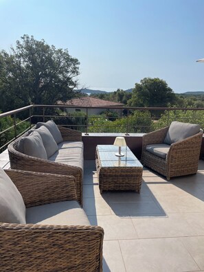 A sunlit outdoor seating area with wicker furniture and a glass-top table, overlooking a scenic landscape.