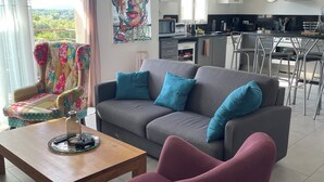 A modern living room with a grey sofa, a wooden coffee table, and a vibrant armchair. A kitchen with a black countertop and bar stools is visible in the background. - Apartment in residence with swimming pool and janitor, walking distance to the beach (Zonza)