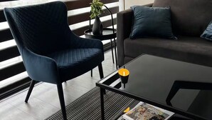 Un salón moderno con un sillón azul oscuro, una mesa de centro negra con una revista, una lámpara de pie negra y un sofá marrón con cojines azules.