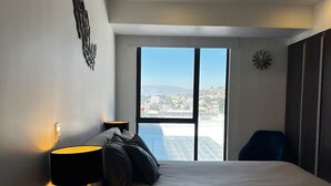 Un dormitorio moderno con una cama grande, un escritorio y una silla. Ofrece vistas de la ciudad a través de amplias ventanas.