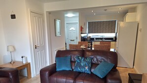 A living room with a brown leather sofa, a wooden side table, and a blue rug. A kitchen is visible in the background.