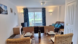 A dining area with a wooden table set for four, surrounded by wicker chairs. A sofa and a television are visible in the background.