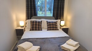 A neatly made bed with a dark blanket and two pillows, two bedside lamps, and a window with curtains.