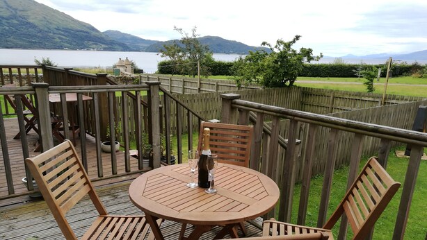 A wooden deck with a round table and chairs, overlooking a lake and mountains.