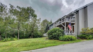 A building with a porch and an American flag, surrounded by trees and a well-maintained lawn. - Cozy Condo on the Mountain (Sugar Mountain)