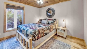 A bedroom with a wooden ceiling, a bed with a patterned quilt, a bedside table with a lamp, and a window with a view of trees. - Cozy Condo on the Mountain (Sugar Mountain)