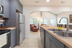 A modern kitchen with grey cabinets, stainless steel appliances, and a wooden countertop. There is a dining area with a chair and a lamp in the background. - Lighthouse Lodge 3 | Lakefront | Sunrise Views + Private Dock | Peaceful Cove (Kimberling City)