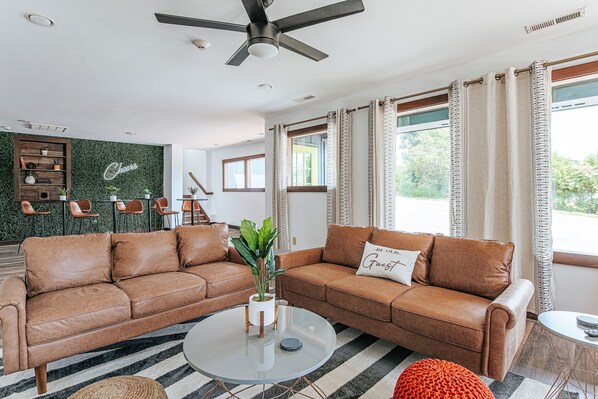 Ein modernes Wohnzimmer mit zwei braunen Ledersofas, einem Glastisch und einer grünen Wand mit einem 'Cheers'-Schild.