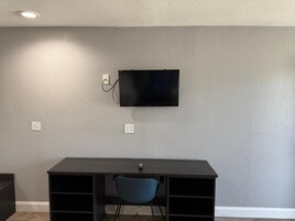 Quarto com escrivaninha de madeira escura, uma cadeira azul e uma TV de tela plana fixada em parede cinza texturizada.