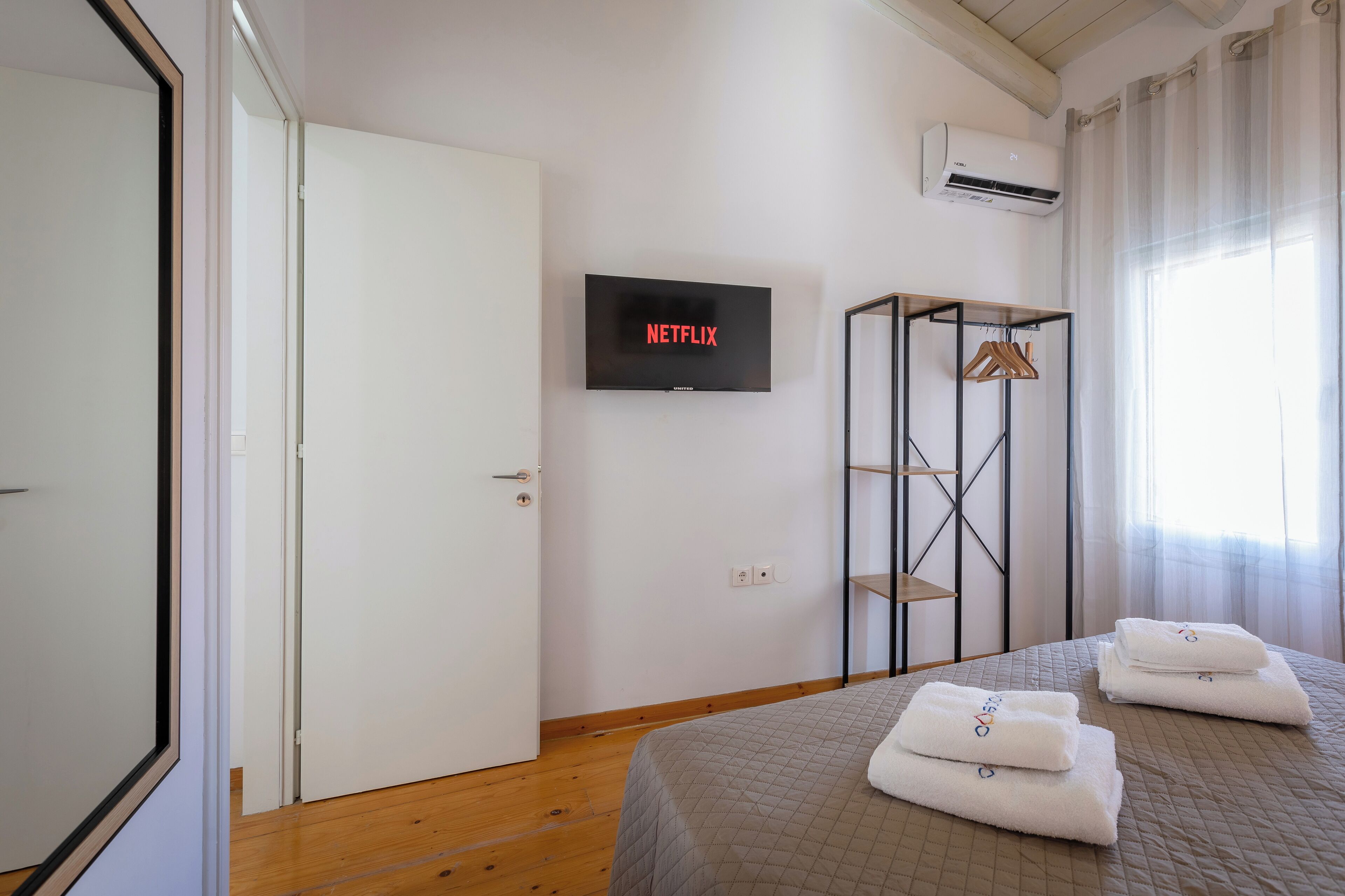 A hotel room with a bed, a TV mounted on the wall, a wardrobe, and an air conditioning unit.