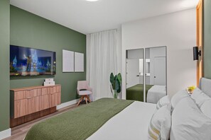 A modern hotel room with a green wall, a large bed, a wooden dresser, a flat-screen TV, a pink chair, and a large mirror.
