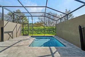 A swimming pool with a glass enclosure, surrounded by a paved area and a hedge.