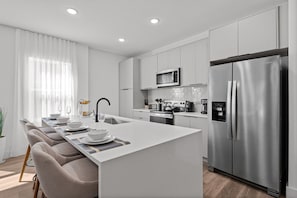A modern kitchen with a central island, stainless steel appliances, and a dining area with chairs and a table set for four. - Perfect Stay Just 15 Min to Disney (Davenport)