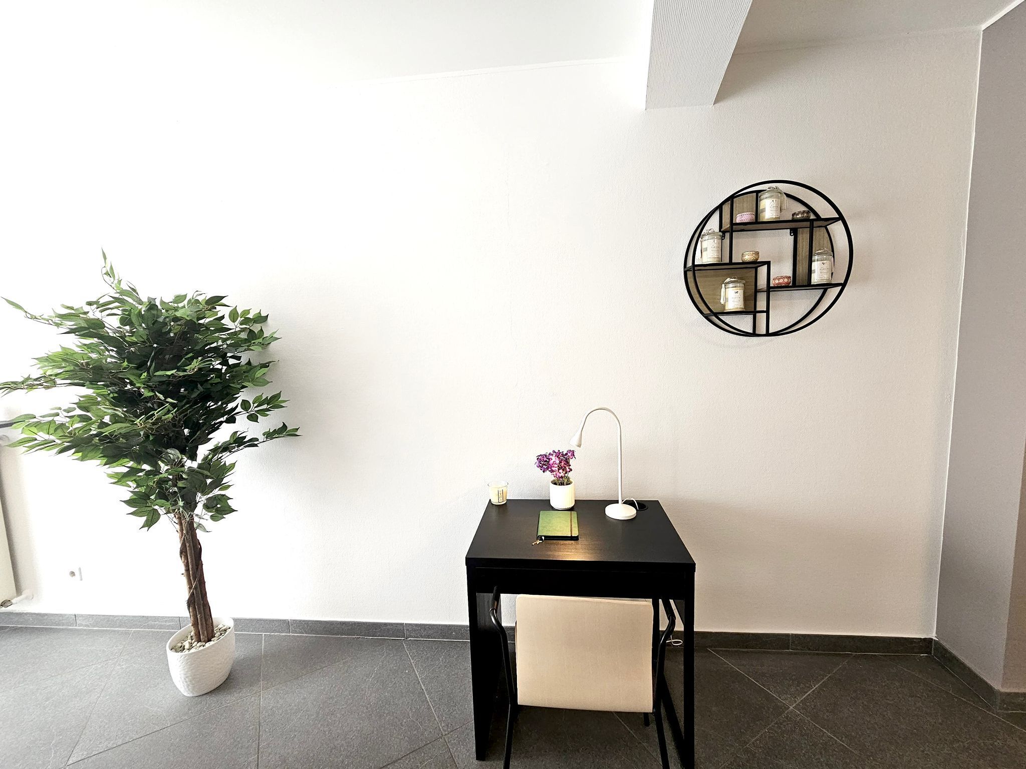 A corner room with a potted plant, a small table with a lamp, and a wall-mounted shelf with jars.