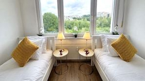 A room with two beds, each with a golden pillow, a small round table with a lamp, and a view of greenery through large windows. - Well Appointed 2BR w terrace&parking (Luxembourg City)