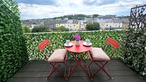 A balcony with a red table and chairs, a vase with flowers, and a view of a town. - Well Appointed 2BR w terrace&parking (Luxembourg City)