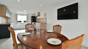 Un comedor con una mesa de madera dispuesta para cuatro personas, rodeado de sillas de mimbre. Se ve una cocina al fondo con un televisor grande montado en la pared.