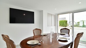 Un comedor con una mesa redonda de madera, cuatro sillas de mimbre, un televisor de pantalla plana montado en la pared y un amplio ventanal con vistas al exterior.