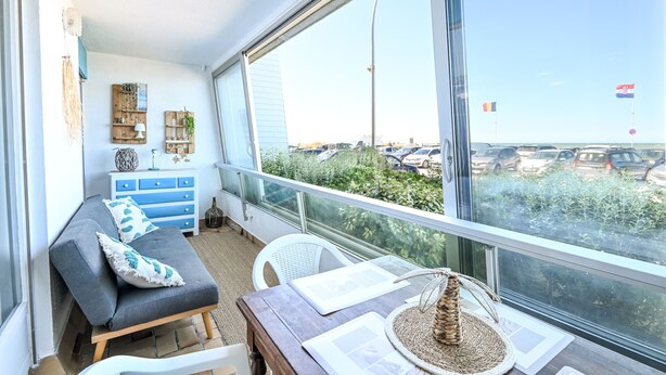 En balkon med sofa, stole, et bord og udsigt til parkering og grøn bevoksning.