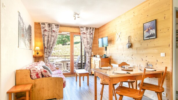 Ein gemütliches Zimmer mit Holzboden, einer Holz-Couch mit gemusterten Kissen, einem Esstisch mit Stühlen, einem an der Wand montierten Fernseher und einem Fenster mit Vorhängen.