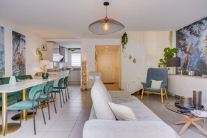 Ein modernes Wohnzimmer mit einer Couch, einem Esstisch und Stühlen. Im Hintergrund ist eine Küche mit Schränken und einem Spülbecken zu sehen.