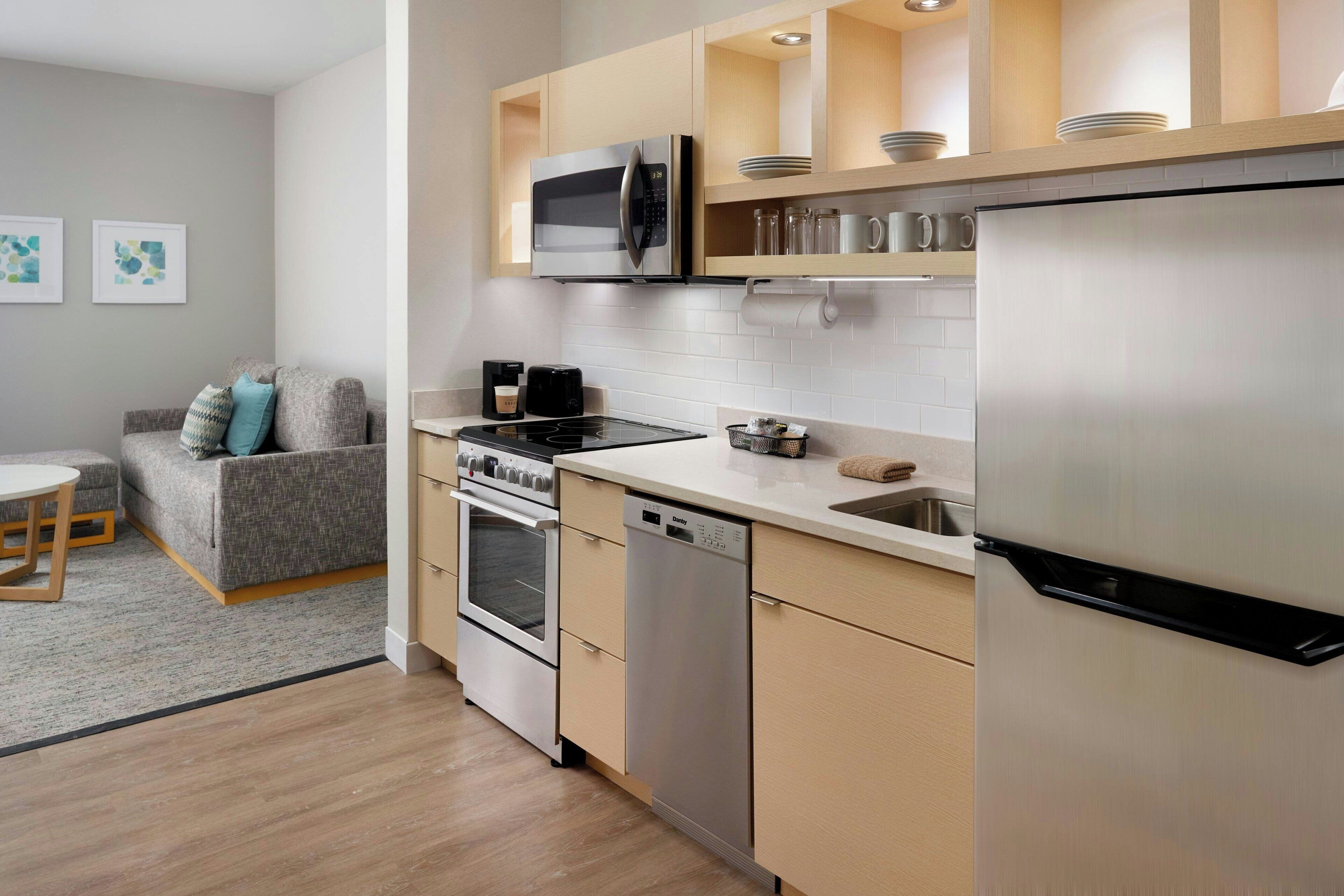 A modern kitchen with stainless steel appliances, a dishwasher, and a built-in oven. There is a dining area with a table and a sofa.
