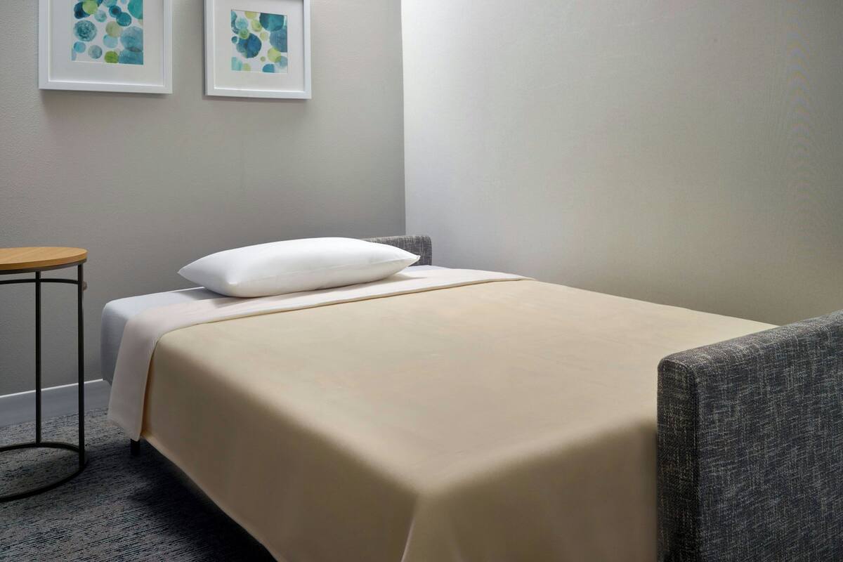 Una cama bien hecha con un almohadón blanco y una colcha color beige en una habitación con un sillón gris y una mesita redonda.