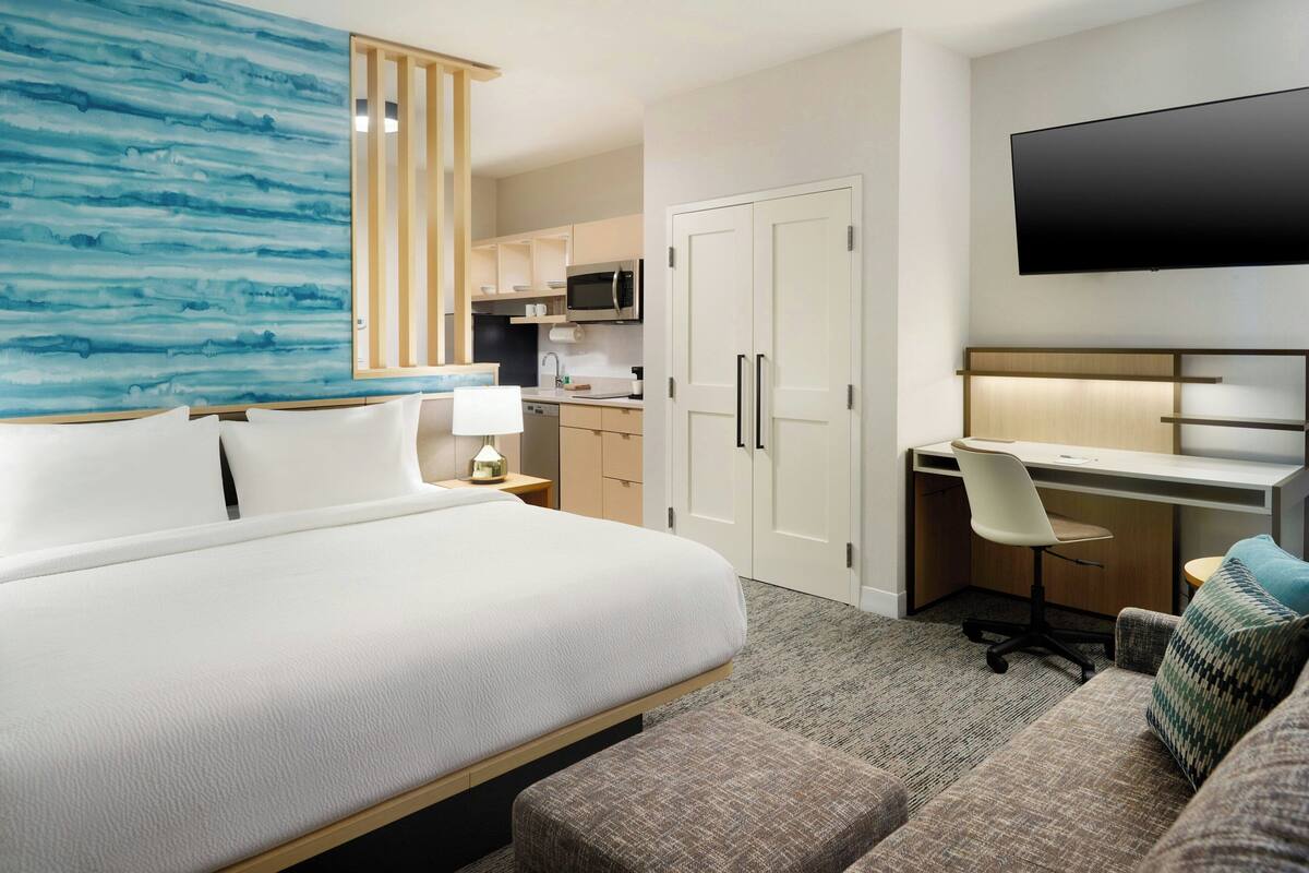 Habitación de hotel con una cama grande, un escritorio con silla, televisor montado en la pared, microondas y kitchenette.