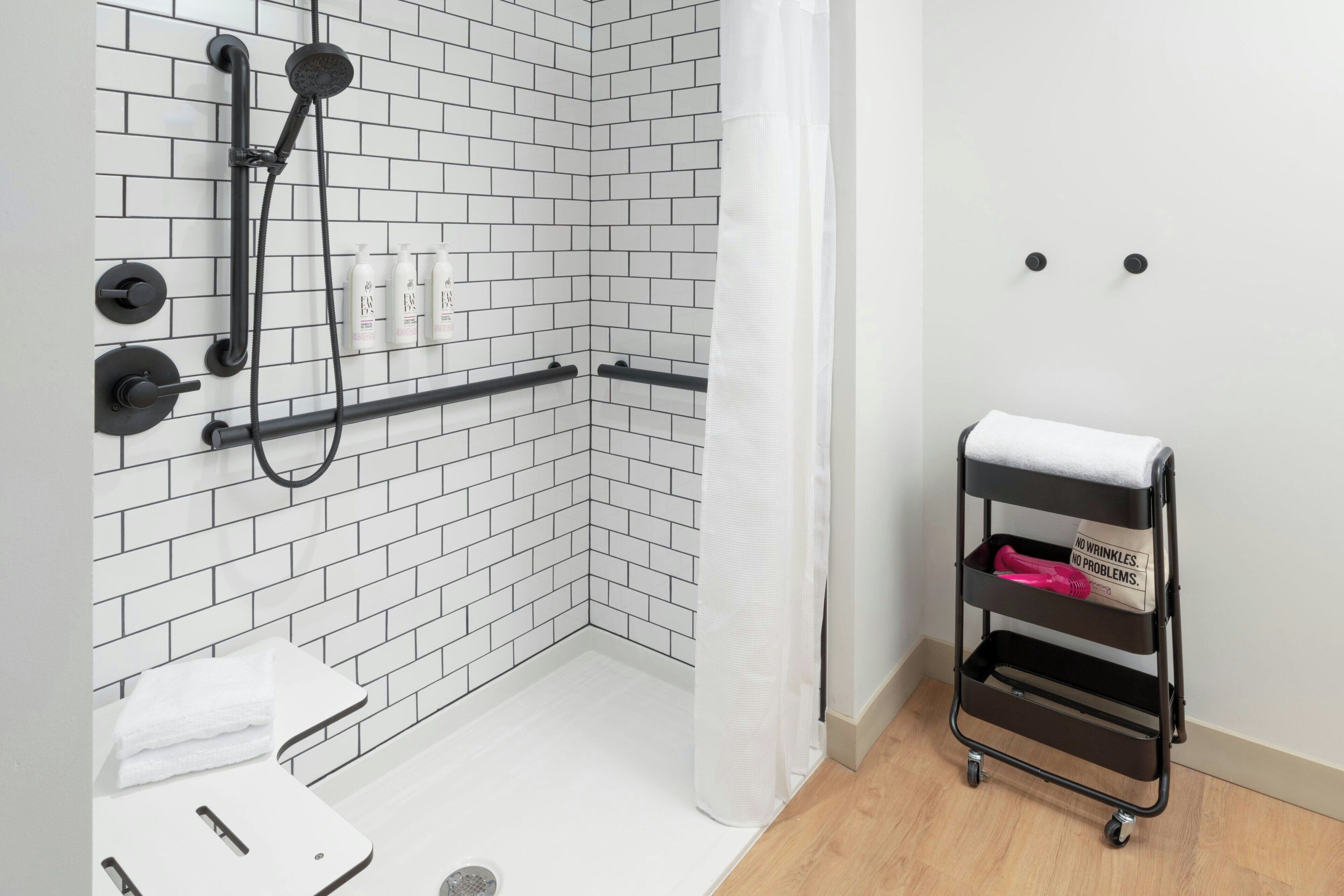 Une salle de bain moderne avec une douche carrelée de carreaux blancs, des toilettes blanches et un chariot de rangement noir avec une serviette.