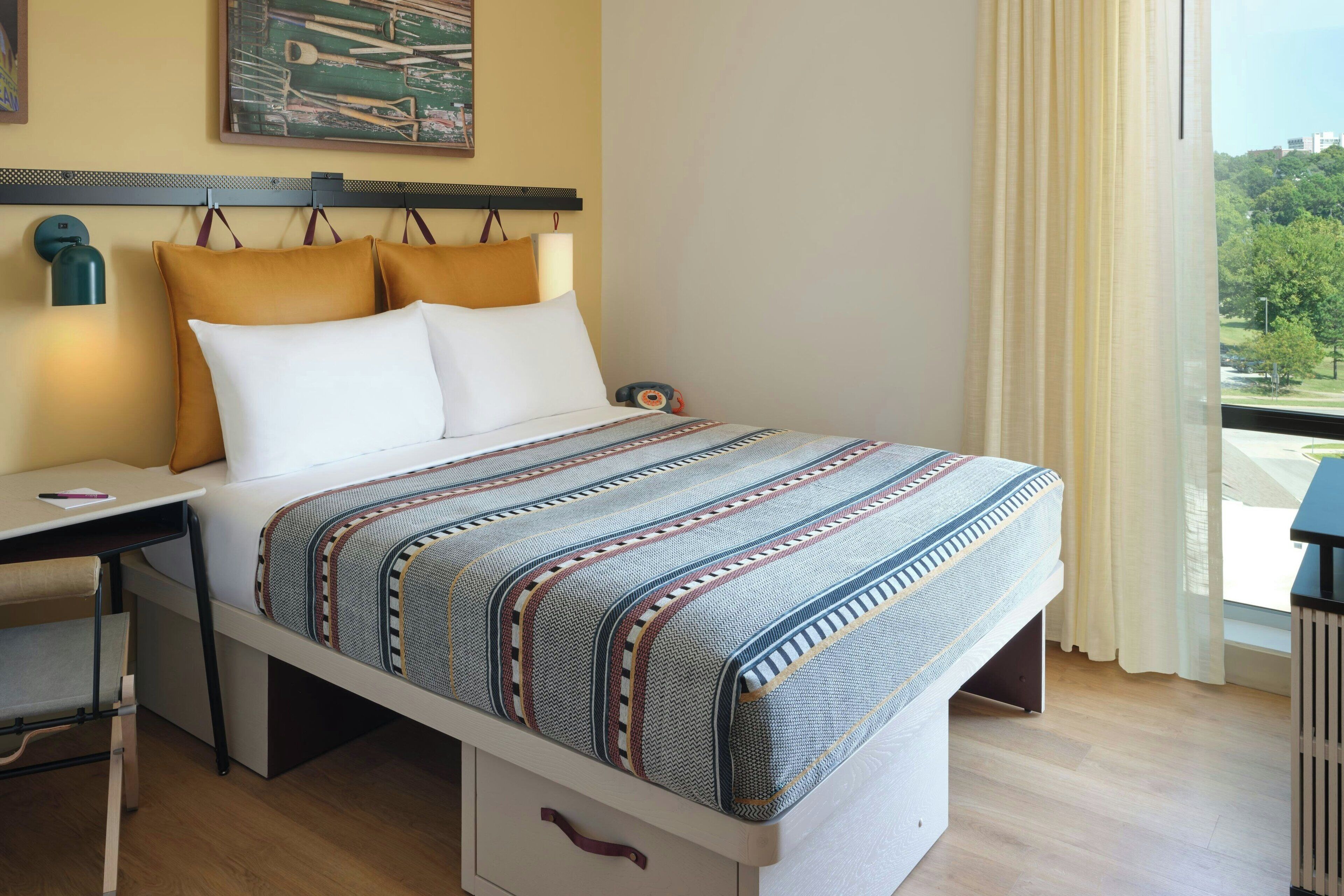 A compact bed with a patterned blanket, a desk with a chair, and a view of greenery through the window.