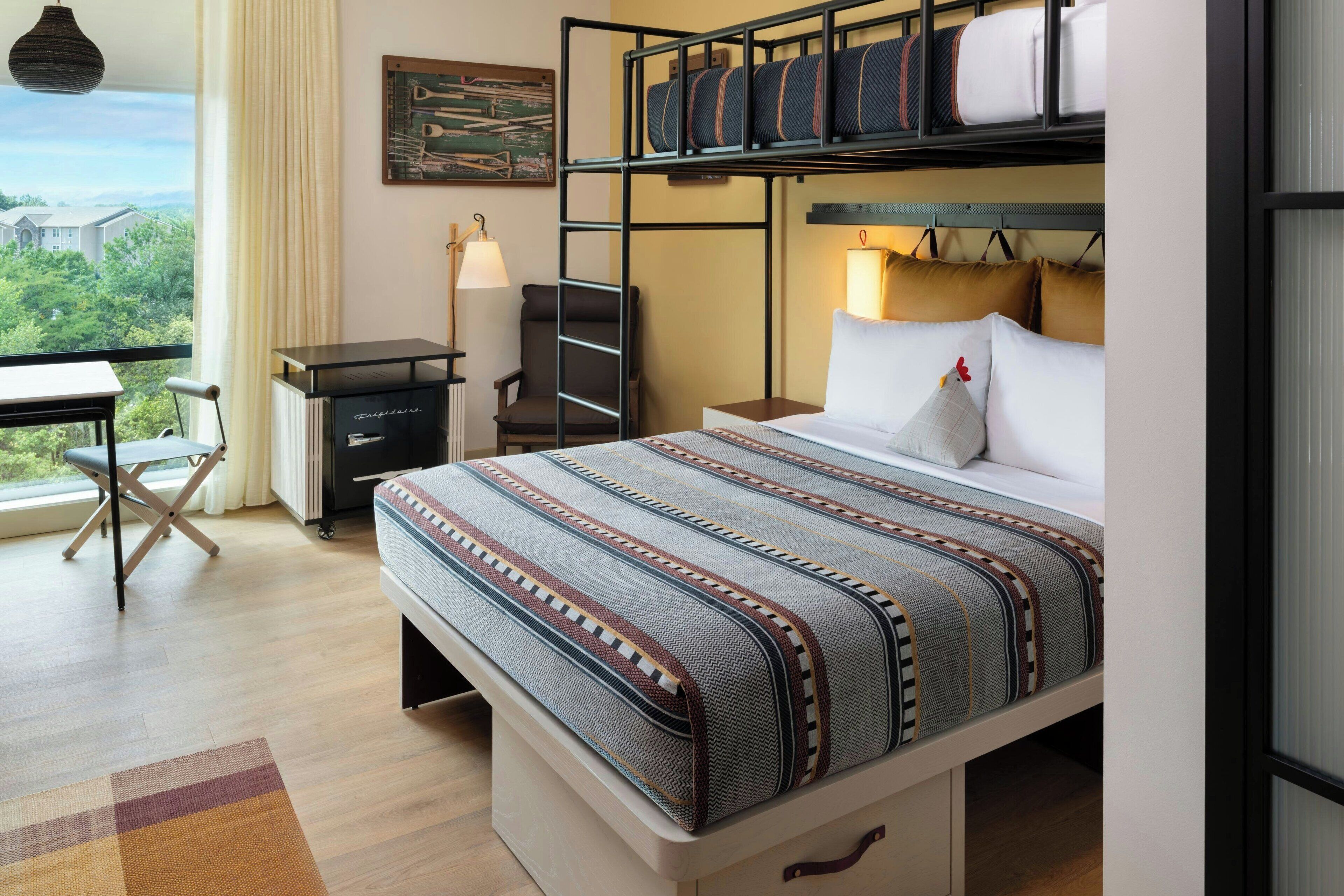 Habitación de hotel con literas, un escritorio con lámpara, una silla y vistas a zonas verdes.