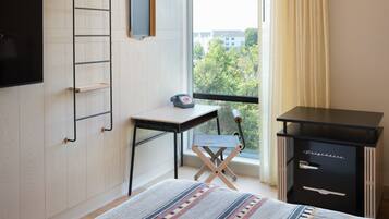 A bedroom with a bed, a desk, a chair, a wardrobe, and a window with a view of buildings and trees.