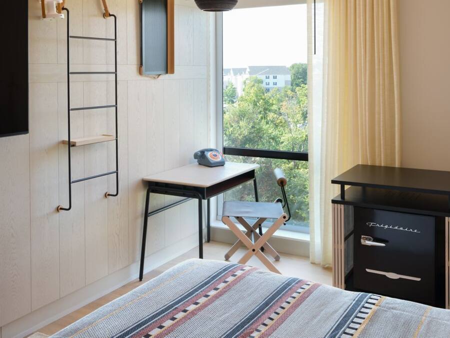 A bedroom with a bed, a desk, a chair, a wardrobe, and a window with a view of buildings and trees.