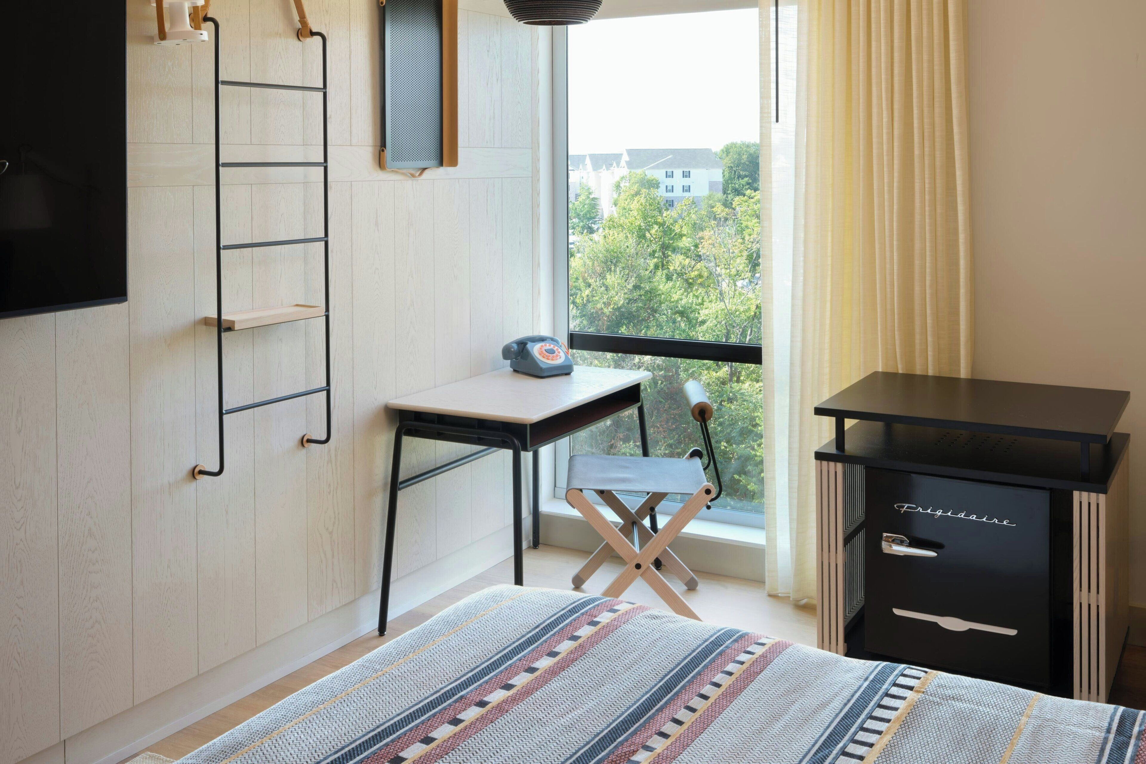 A bedroom with a bed, a desk, a chair, a wardrobe, and a window with a view of buildings and trees.