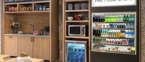 A snack bar with a microwave, refrigerator, and shelves stocked with snacks and beverages.