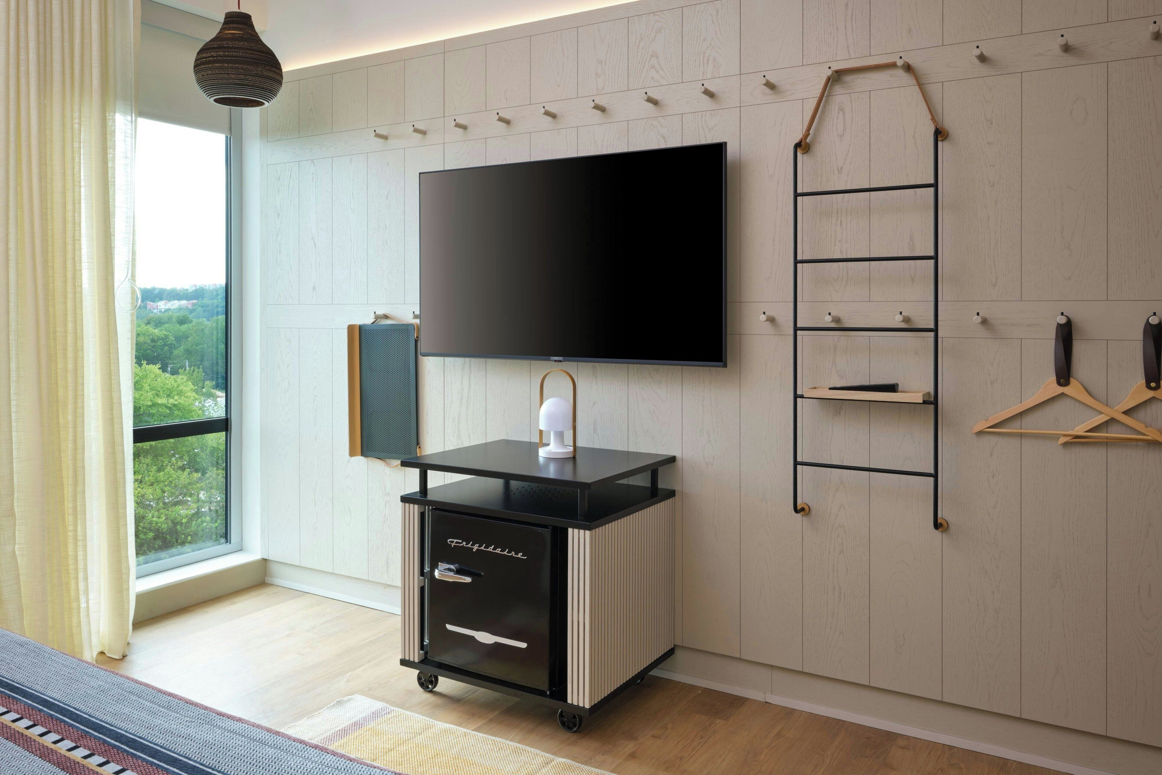 A modern hotel room with a large TV mounted on the wall, a black and white minibar, a wooden ladder, and a view of greenery through the window.