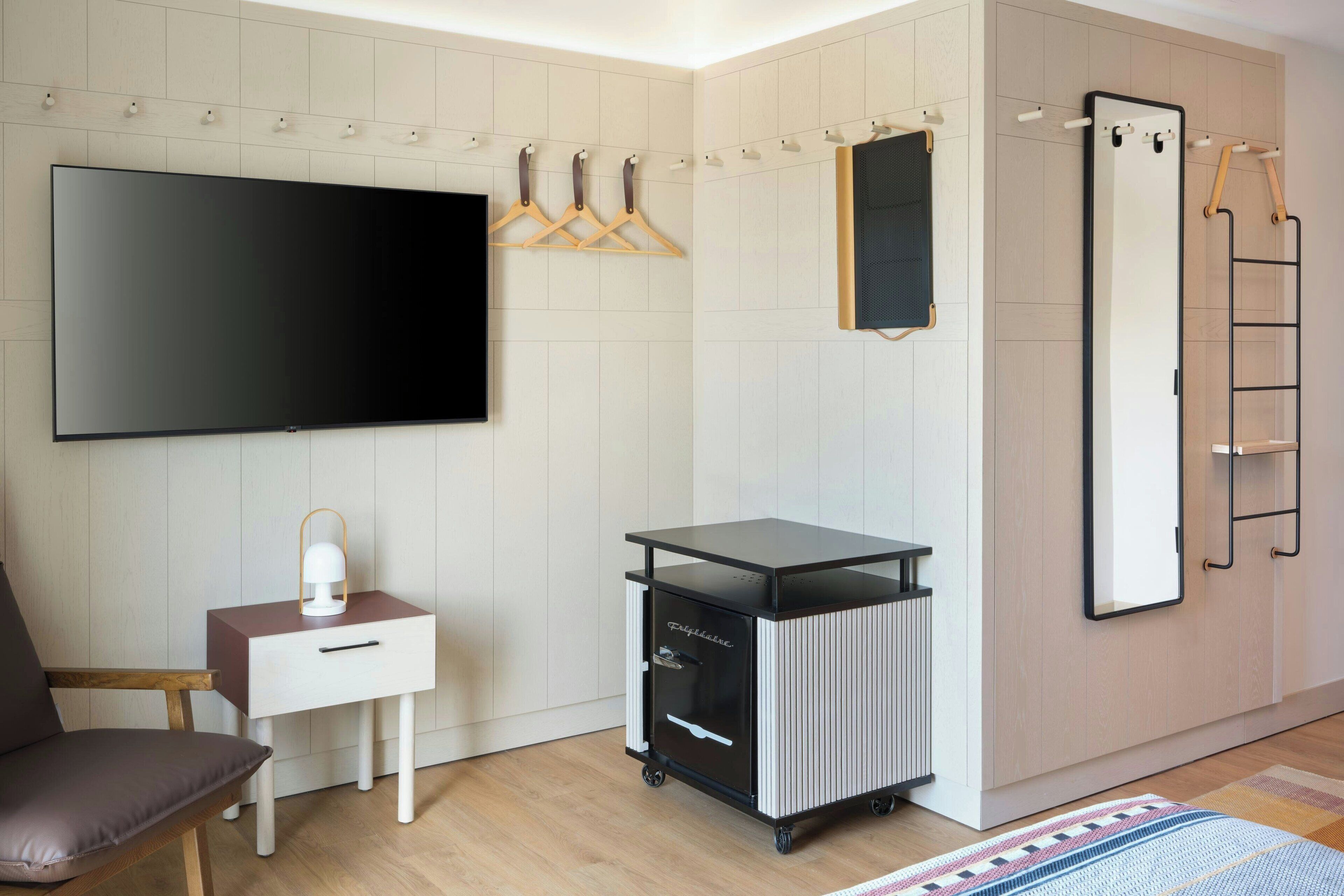 Ett modernt hotellrum med en platt-TV, ett litet skrivbord med en lampa, en förvaringsvagn, en garderob och en spegel.