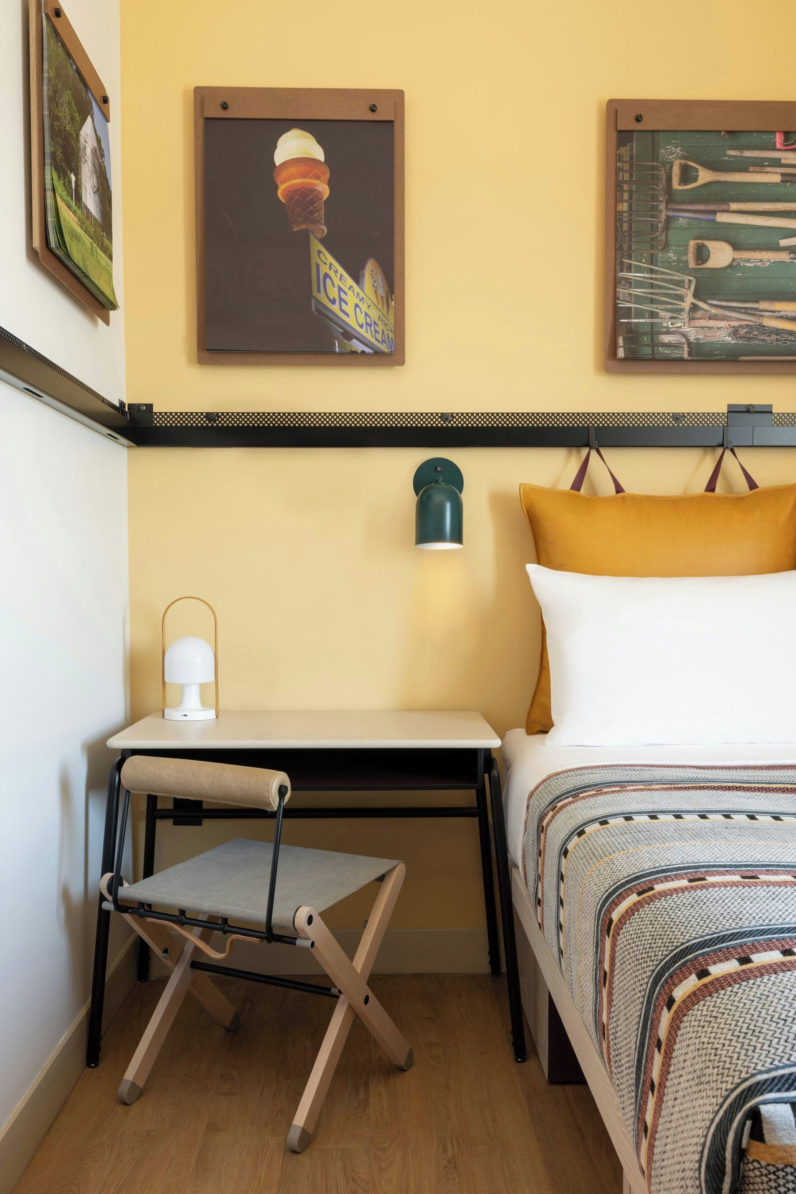 A compact hotel room with a bed, a small desk, a chair, a nightstand, a lamp, and two wall art pieces.