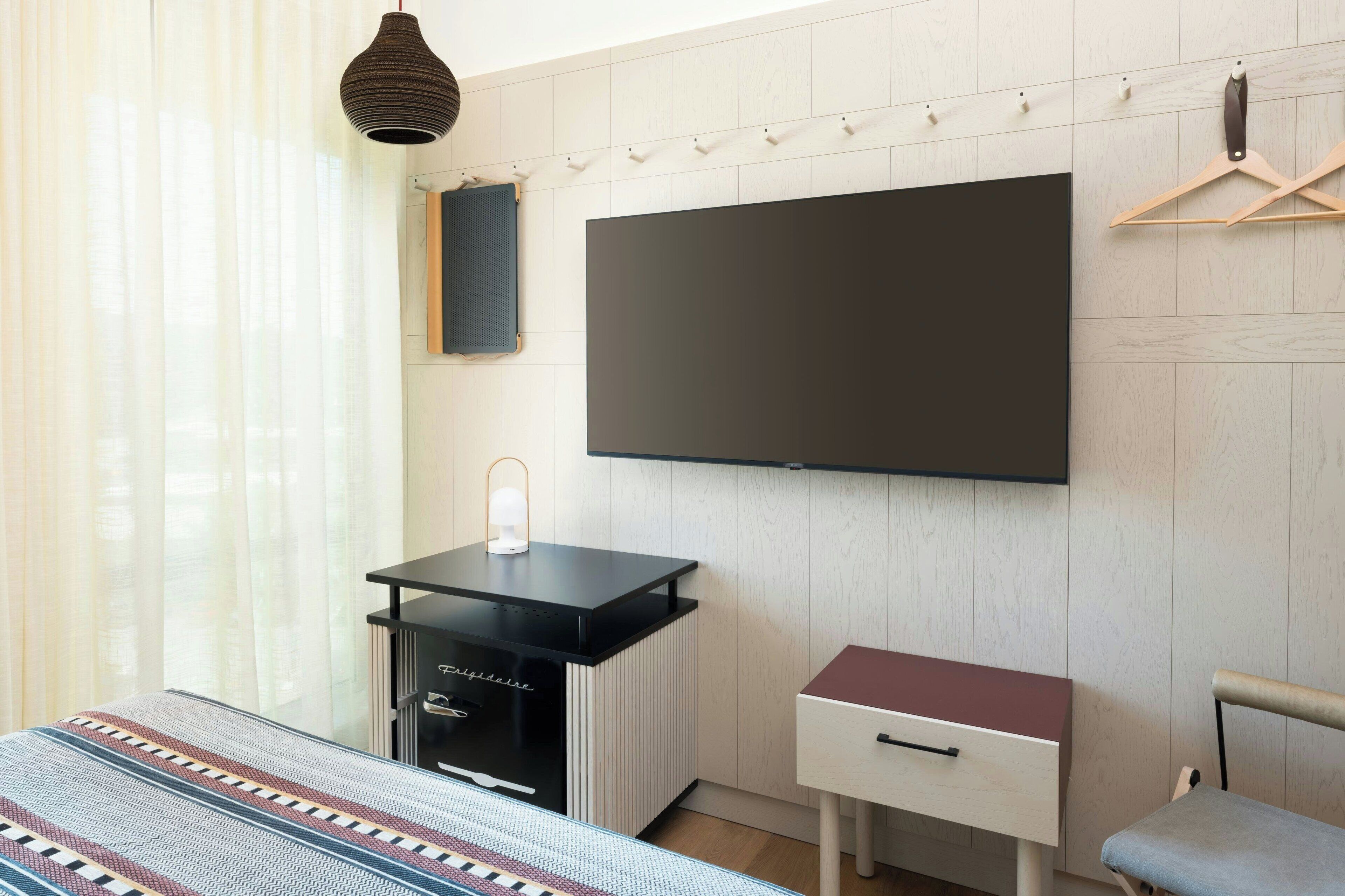 Quarto de hotel moderno com uma grande TV de tela plana na parede, uma pequena escrivaninha preta com luminária, mesa de cabeceira branca e uma cadeira cinza.