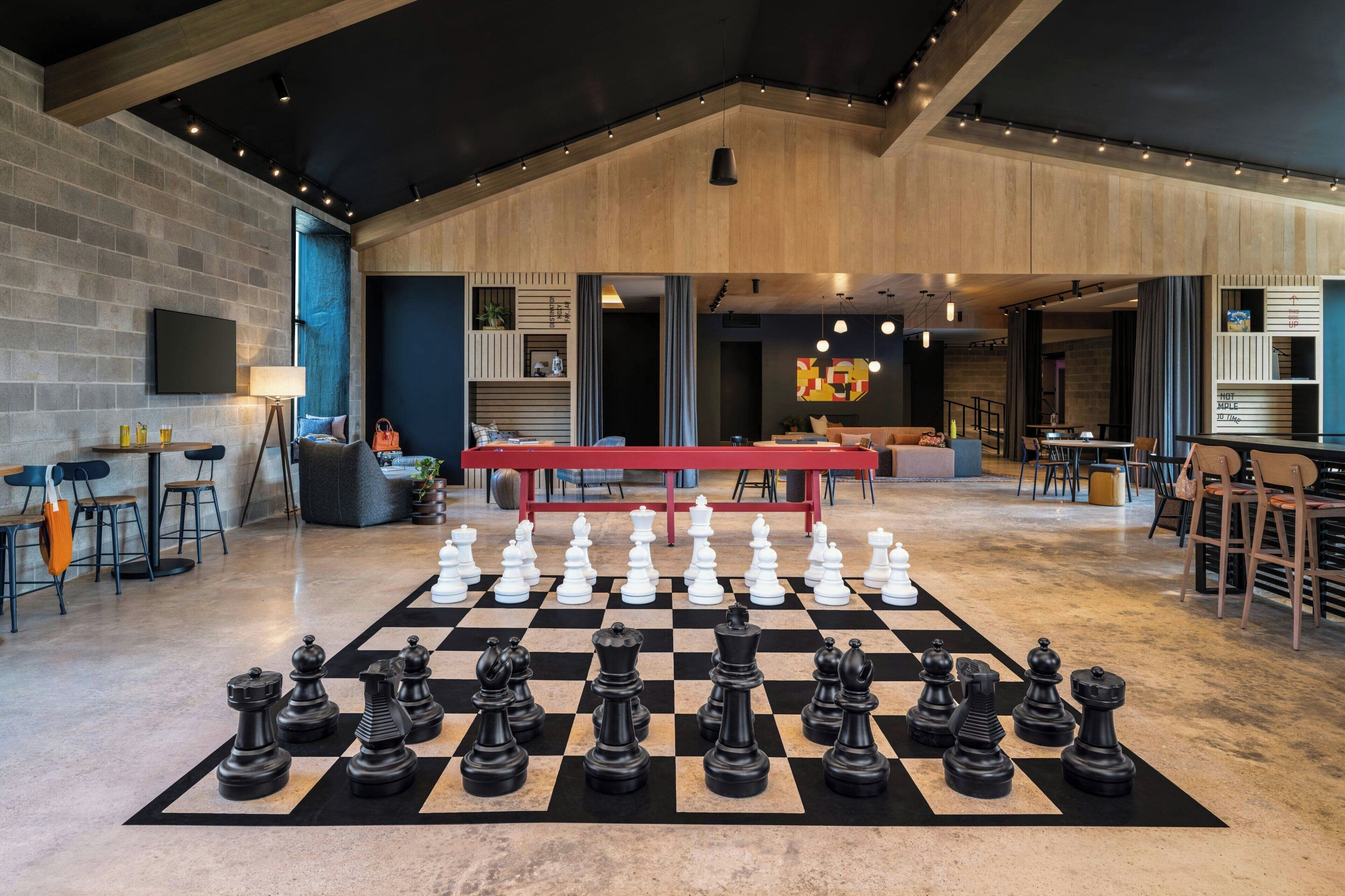 A spacious room with a large chessboard set up in the center, surrounded by modern furniture and a high ceiling with exposed beams.