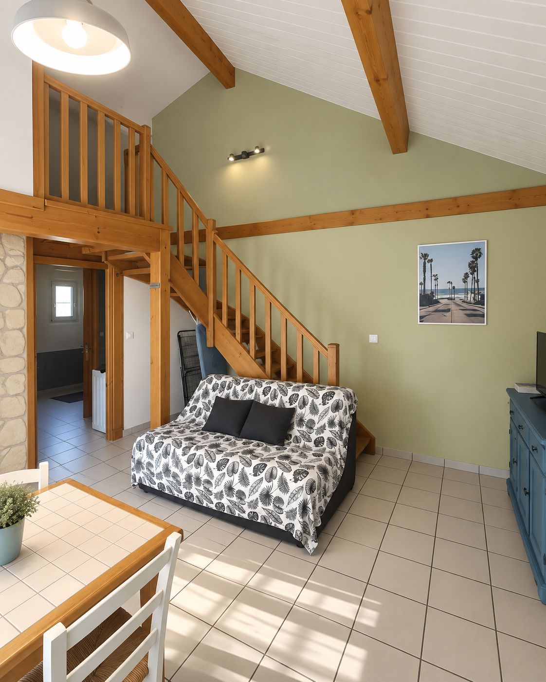 Un salon chaleureux avec un escalier en bois, un canapé orné d’un motif végétal, une table à manger et un tableau représentant une scène de plage.