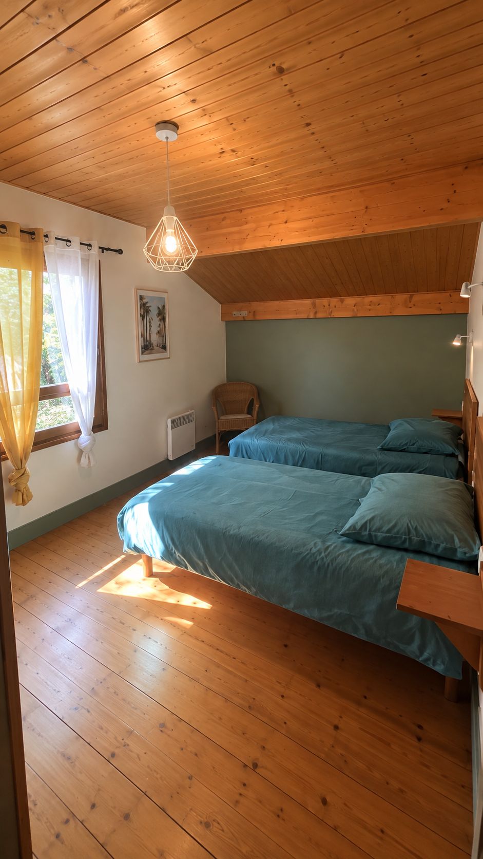 Een kamer met houten vloeren, een bed met blauw beddengoed, een hanglamp en een raam met gordijnen.