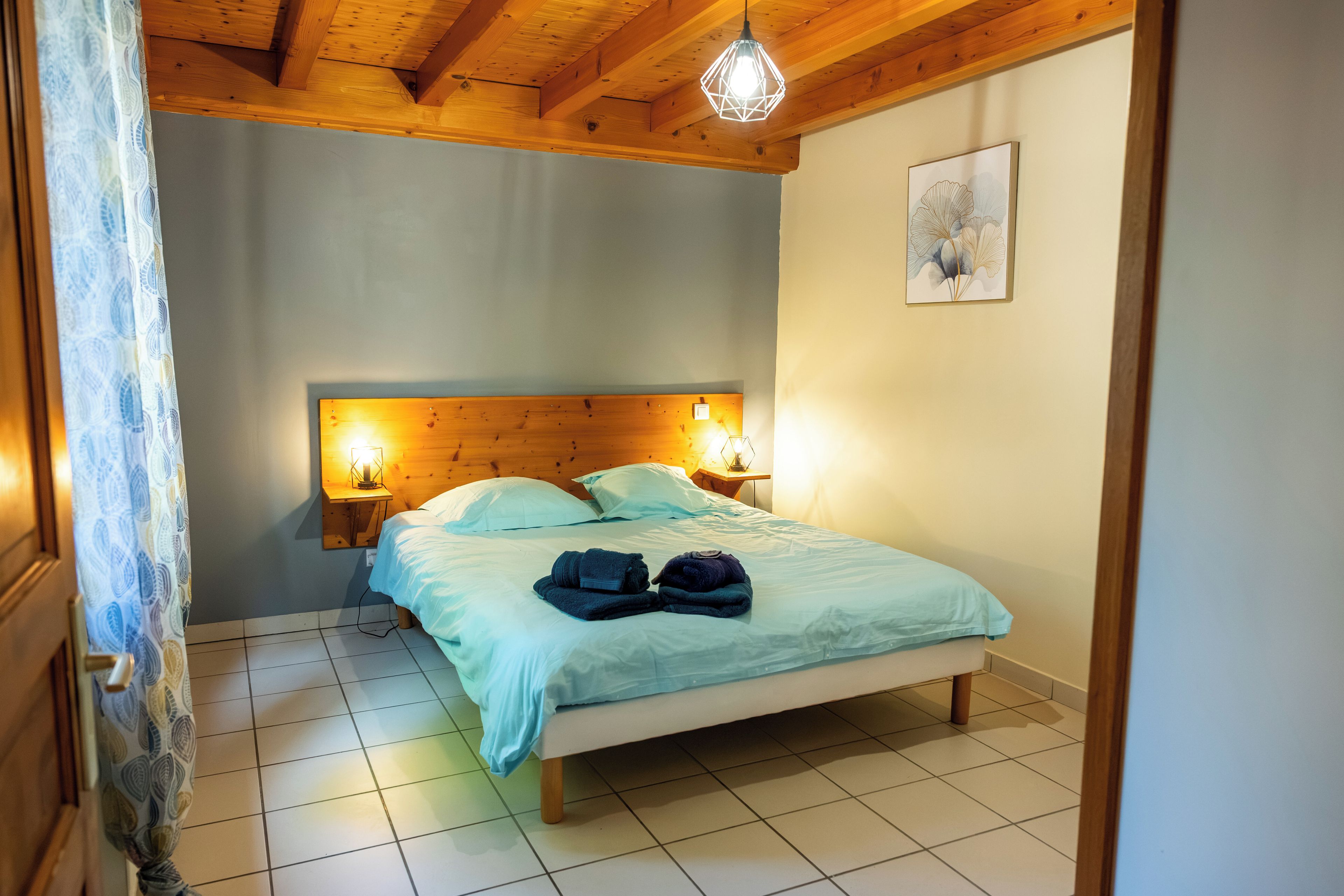Un dormitorio con techo de madera, una cama con colcha azul, dos lámparas de noche y una obra de arte colgada en la pared.