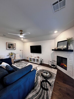 Ein modernes Wohnzimmer mit einem blauen Sofa, einem Kamin, einem Wand-Fernseher und einem Deckenventilator.