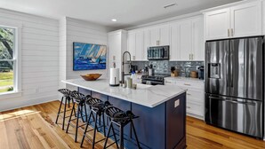 Une cuisine moderne avec des armoires blanches, des appareils en acier inoxydable, un grand îlot central doté de tabourets de bar et un tableau représentant un voilier accroché au mur.