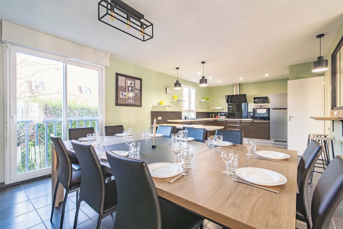 Uma sala de jantar com uma mesa grande posta para seis pessoas, uma cozinha ao fundo e uma varanda com vista.