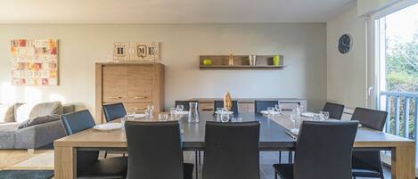 A modern dining area with a wooden table and black chairs, a kitchen area with cabinets, and a view of the outdoors through a large window.