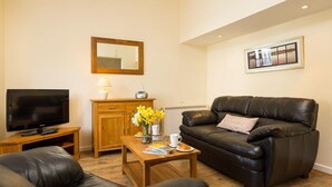 A living room with a leather sofa, a wooden coffee table with a vase of flowers, a television, and a cabinet. - 2 Riverside Cottage (Ambleside)