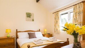 A bedroom with a wooden bed, two bedside tables with lamps, a window with floral curtains, and a vase with flowers on a dresser. - 2 Riverside Cottage (Ambleside)